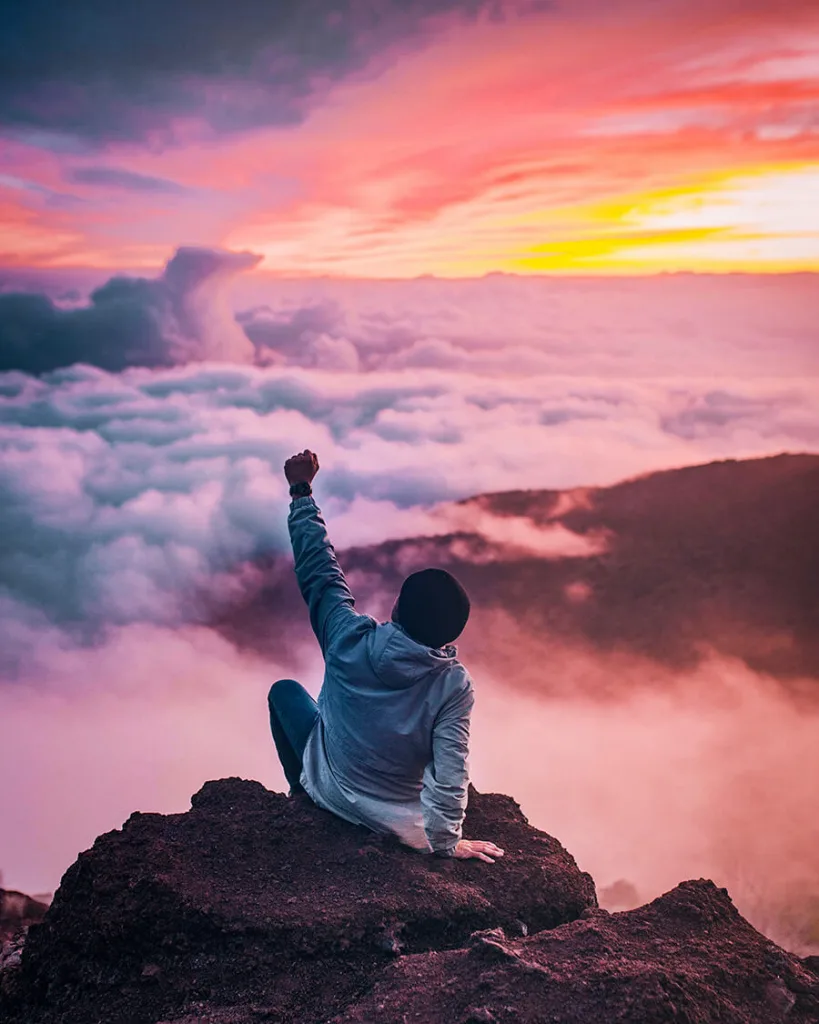 Person sitting on a mountain top above the clouds at sunrise with one arm raised